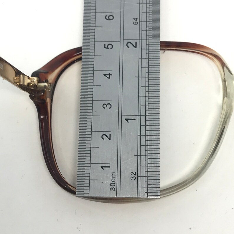 Op de afbeelding: Close-up van een bril met bruine monturen en heldere glazen. Een metalen liniaal is over de bril geplaatst, met een breedte van ongeveer 5 cm. De bril ligt op een witte ondergrond.