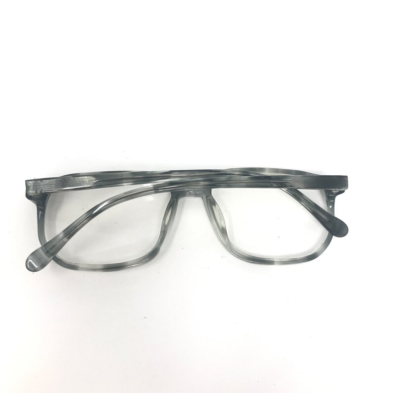 Puede incluir: Gafas grises y blancas veteadas con montura rectangular y lentes transparentes. Las gafas tienen un dise&ntilde;o cl&aacute;sico con una forma ligeramente sobredimensionada. Las patillas son finas y se extienden hasta los terminales.