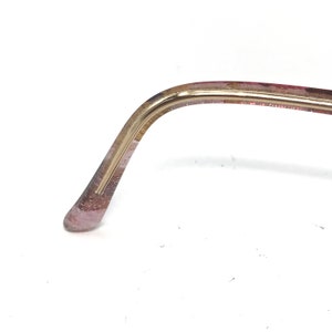 May include: Close-up of a portion of eyeglasses. The frame is a combination of gold and translucent pink. The gold portion curves to meet the pink, which has a glittery appearance. The glasses are set against a white background.