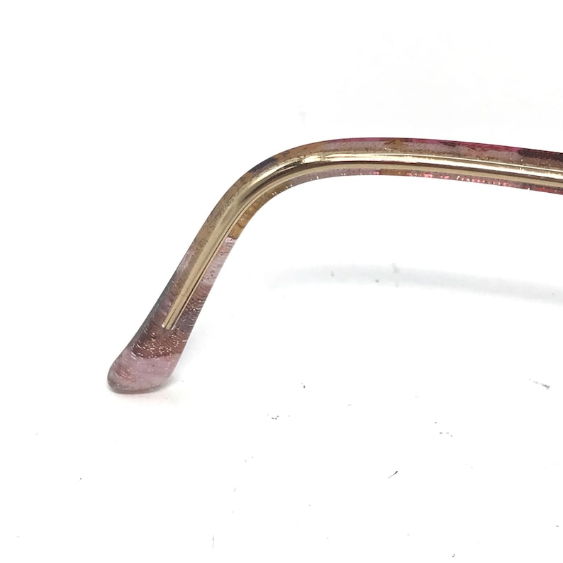 May include: Close-up of a portion of eyeglasses. The frame is a combination of gold and translucent pink. The gold portion curves to meet the pink, which has a glittery appearance. The glasses are set against a white background.