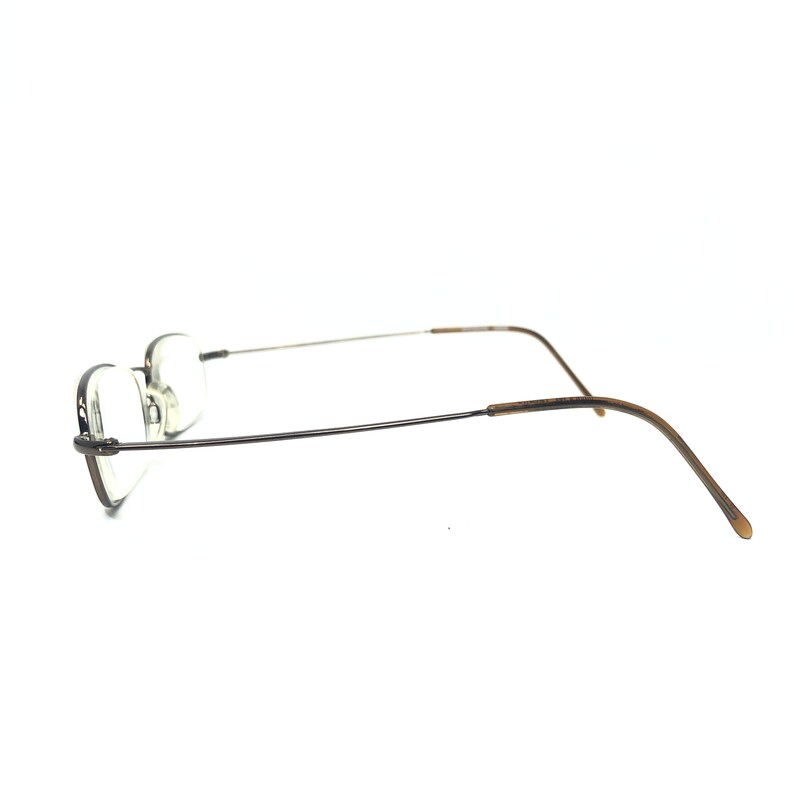 Puede incluir: Un par de gafas sin montura con forma de lente rectangular. Las monturas son de color met&aacute;lico oscuro, con varillas finas plateadas y patillas marrones. Las gafas est&aacute;n sobre un fondo blanco.