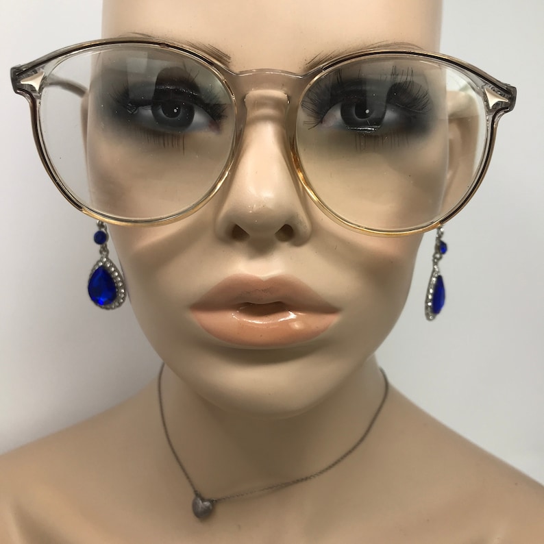Puede incluir: Gafas transparentes con montura dorada y puente transparente. Las gafas est&aacute;n sobre una cabeza de maniqu&iacute;. El maniqu&iacute; lleva pendientes azules en forma de l&aacute;grima con detalles plateados y un collar con colgante de coraz&oacute;n plateado.