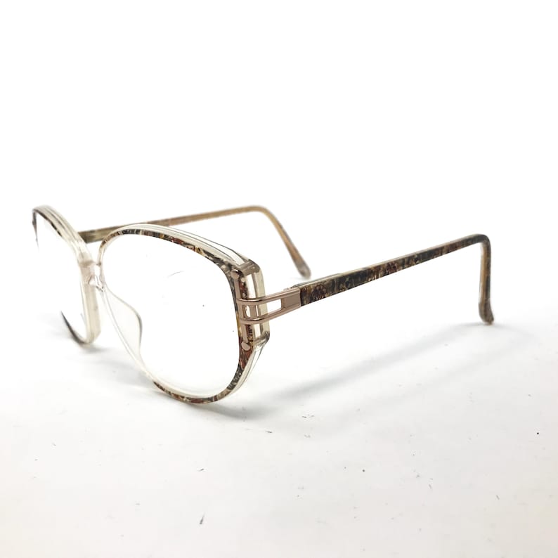 May include: A pair of brown and gold eyeglasses with a patterned frame. The frame is made of plastic and has a clear lens.