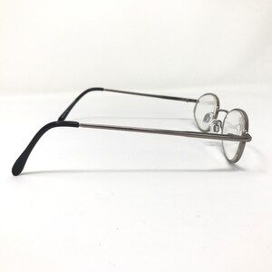 Op de afbeelding: Een bril met een dun, donkergrijs metalen montuur en ronde glazen. De pootjes zijn zwart en buigen naar beneden om achter de oren te rusten. De bril staat tegen een witte achtergrond.