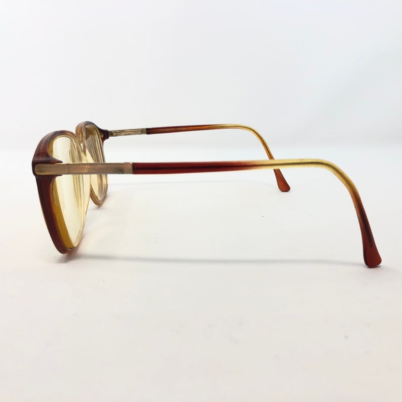 May include: A pair of vintage eyeglasses with a brown and gold gradient frame. The lenses are a light yellow color. The glasses have a rectangular shape and are viewed from the side.