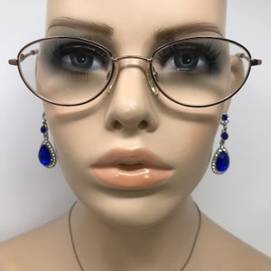 Op de afbeelding: Een mannequin draagt ovale bril met een ros&eacute;gouden montuur en blauwe traanvormige oorbellen met strass.