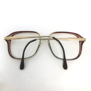 Op de afbeelding: Vintage bril met een bruin en goudkleurig montuur. De lenzen zijn helder en de pootjes zijn zwart. De bril heeft een rechthoekige vorm en een gouden brug. De algehele stijl is retro.