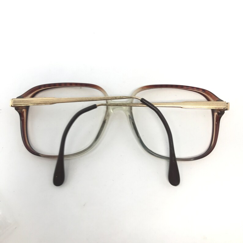 Op de afbeelding: Vintage bril met een bruin en goudkleurig montuur. De lenzen zijn helder en de pootjes zijn zwart. De bril heeft een rechthoekige vorm en een gouden brug. De algehele stijl is retro.
