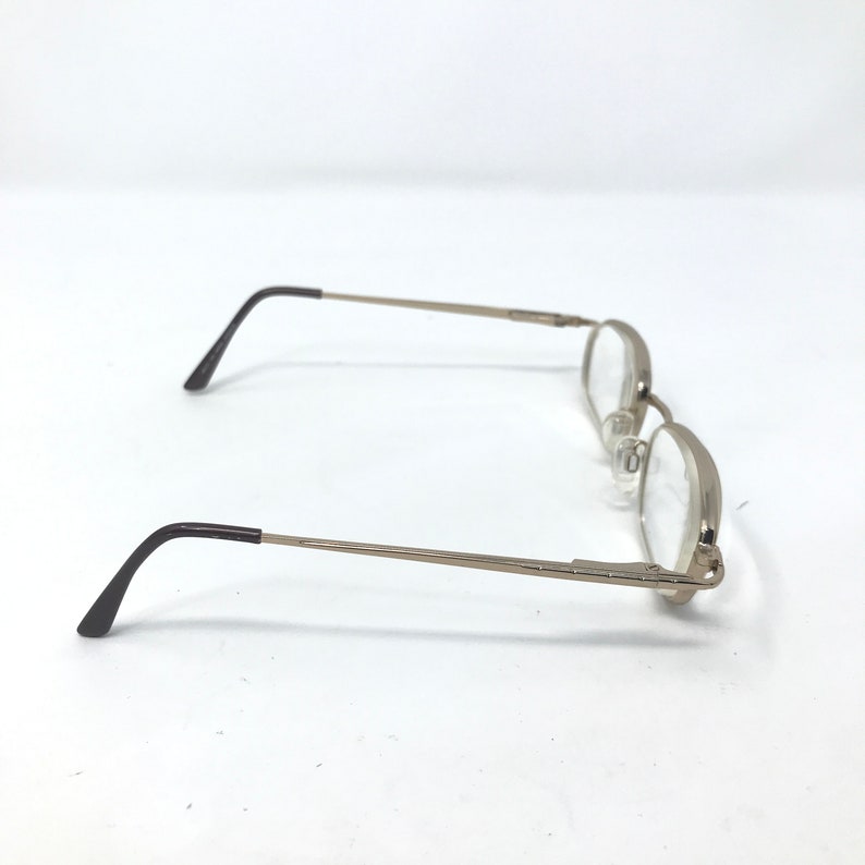 May include: Silver-toned eyeglasses with a rectangular frame and clear lenses. The temples are thin and extend to black tips. The glasses are set against a white background.