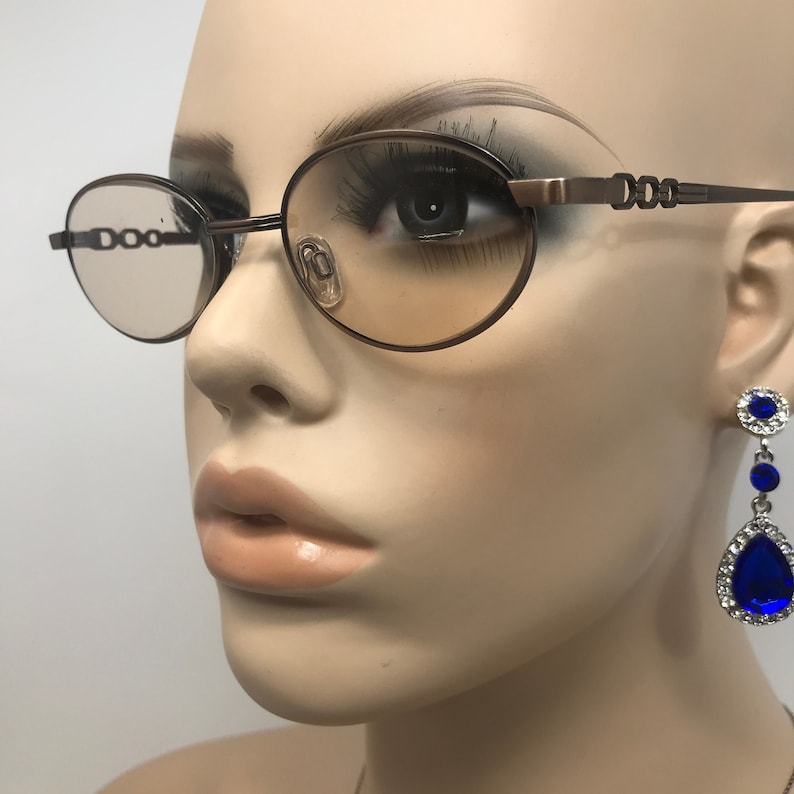 May include: Brown oval-shaped eyeglasses with a decorative chain design on the temples. The lenses have a light tint. A blue and silver teardrop earring is visible on the right side of the image. The glasses are on a mannequin.