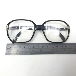 May include: Black-framed eyeglasses with clear lenses. The glasses have a rectangular shape with rounded edges and silver accents on the temples. A ruler is placed below the glasses, measuring approximately 15 cm in length.