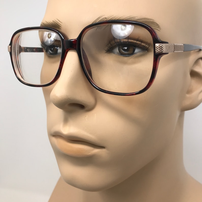 May include: Square-framed eyeglasses with a tortoiseshell pattern on the rims and temples. The glasses have clear lenses and gold-colored accents on the temples. The frames are set on a mannequin head.