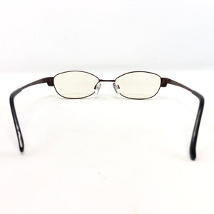 May include: A pair of oval-shaped eyeglasses with a dark brown metal frame. The lenses are clear, and the temples are black. The glasses are set against a white background.