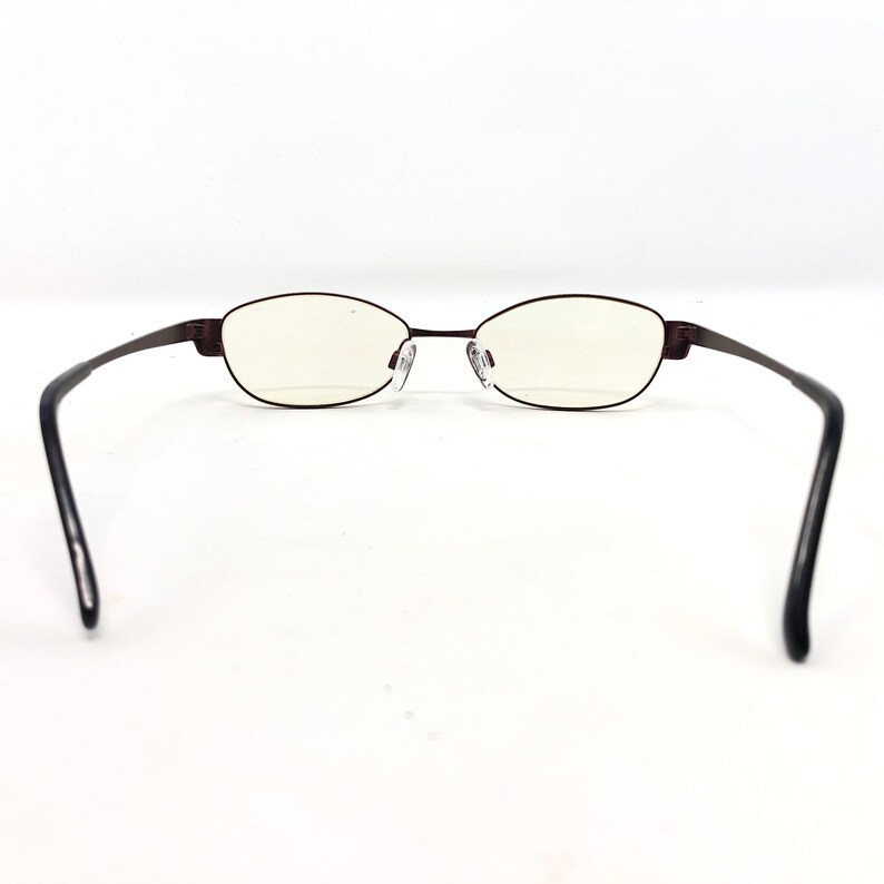 May include: A pair of oval-shaped eyeglasses with a dark brown metal frame. The lenses are clear, and the temples are black. The glasses are set against a white background.