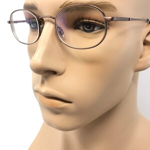 Puede incluir: Primer plano de gafas con montura met&aacute;lica fina. Las lentes son redondas y la montura tiene un color neutro sutil. Las gafas se muestran en una cabeza de maniqu&iacute;, mostrando el dise&ntilde;o y el ajuste.