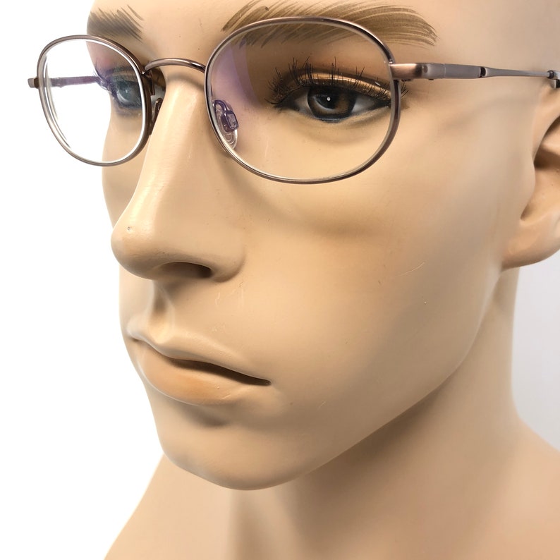 Puede incluir: Primer plano de gafas con montura met&aacute;lica fina. Las lentes son redondas y la montura tiene un color neutro sutil. Las gafas se muestran en una cabeza de maniqu&iacute;, mostrando el dise&ntilde;o y el ajuste.