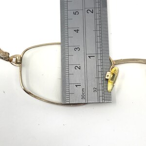 May include: Close-up of gold-rimmed spectacles with clear lenses. A ruler is placed next to the glasses, showing a measurement of approximately 2.5 cm. The spectacles have a classic, rectangular shape.