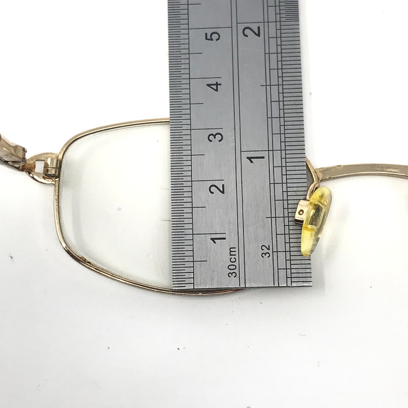 May include: Close-up of gold-rimmed spectacles with clear lenses. A ruler is placed next to the glasses, showing a measurement of approximately 2.5 cm. The spectacles have a classic, rectangular shape.