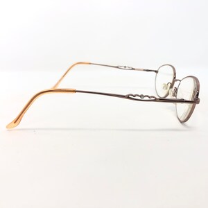 May include: A pair of eyeglasses with a brown metal frame and oval lenses. The temples are a light brown color, transitioning to a darker brown with decorative cutouts. The glasses are set against a white background.