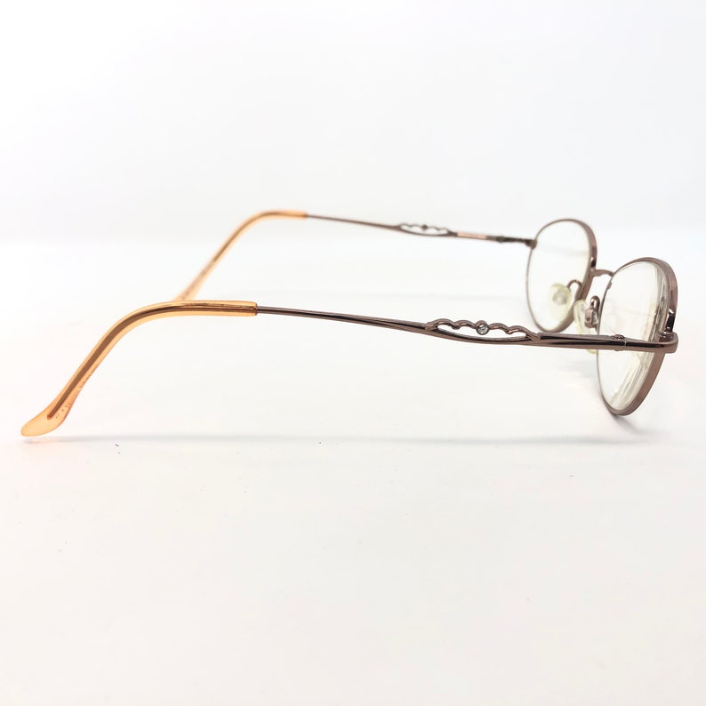 May include: A pair of eyeglasses with a brown metal frame and oval lenses. The temples are a light brown color, transitioning to a darker brown with decorative cutouts. The glasses are set against a white background.