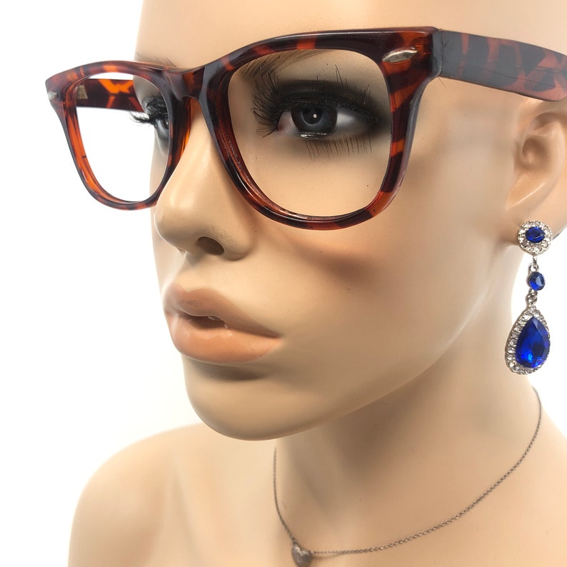 K&ouml;nnte beinhalten: Brille mit quadratischem Rahmen und Schildpattmuster. Die Brille befindet sich auf einem Schaufensterkopf. Die Schaufensterpuppe tr&auml;gt au&szlig;erdem ein Paar blaue Tropfenohrringe mit silberfarbener Fassung und eine silberne Halskette mit Herzanh&auml;nger.