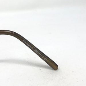 May include: Close-up of the arm of a pair of eyeglasses. The arm is a dark brown color and has the text "Specsavers 1003314+ L CE" printed on it in white. The background is plain white.