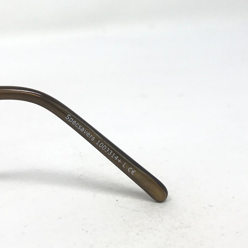 May include: Close-up of the arm of a pair of eyeglasses. The arm is a dark brown color and has the text "Specsavers 1003314+ L CE" printed on it in white. The background is plain white.
