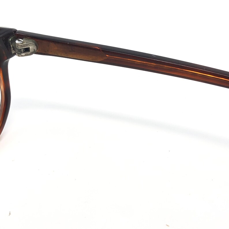 May include: Close-up of a pair of brown eyeglasses. The frame is a dark brown color with a rectangular shape. The arm of the glasses is extended, showing the hinge and the sleek design. The background is white.