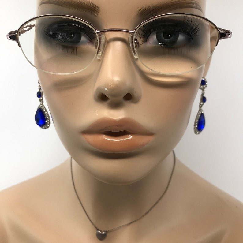 K&ouml;nnte beinhalten: Eine Mannequin tr&auml;gt eine silberne Halskette mit einem Herzanh&auml;nger und eine ovale Brille mit einem ros&eacute;goldenen Gestell. Die Mannequin tr&auml;gt auch ein Paar Ohrringe mit blauen, tropfenf&ouml;rmigen Steinen.