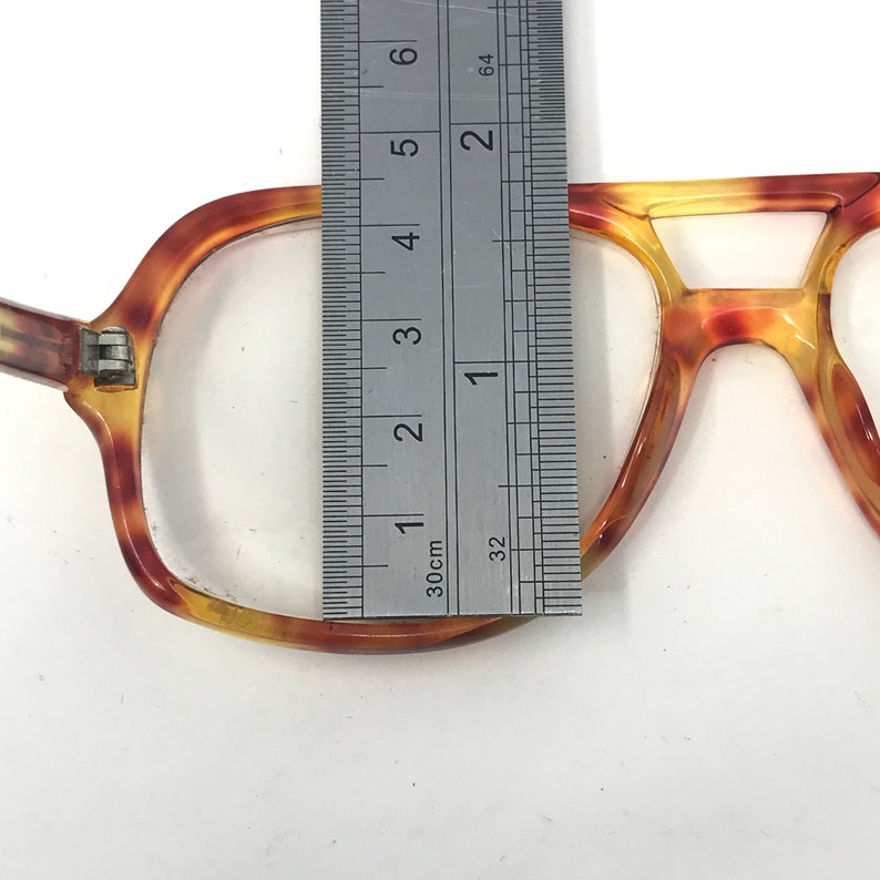 May include: Close-up of vintage eyeglasses with a tortoise shell pattern. The frame is measured with a ruler, showing a width of approximately 1 inch. The glasses have a classic, rectangular shape.