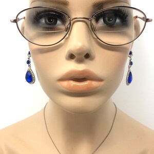 May include: A mannequin wearing oval-shaped eyeglasses with a thin, rose-gold frame. Dangling earrings with large, blue teardrop-shaped gems and smaller blue gems are visible. A silver necklace is also present. The mannequin has full lips and is set against a white background.