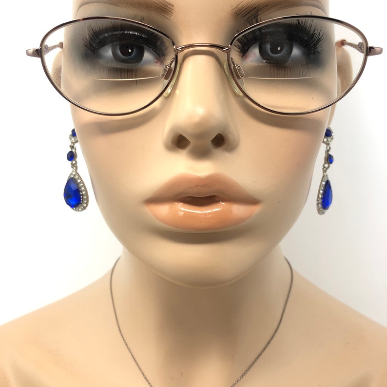 May include: A mannequin wearing oval-shaped eyeglasses with a thin, rose-gold frame. Dangling earrings with large, blue teardrop-shaped gems and smaller blue gems are visible. A silver necklace is also present. The mannequin has full lips and is set against a white background.