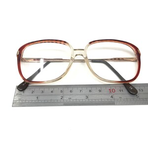 K&ouml;nnte beinhalten: Braune und klare Kunststoff-Brille mit einem Metallsteg und B&uuml;geln. Die Brille wird mit einem Lineal gemessen und zeigt eine Breite von etwa 10 Zentimetern.