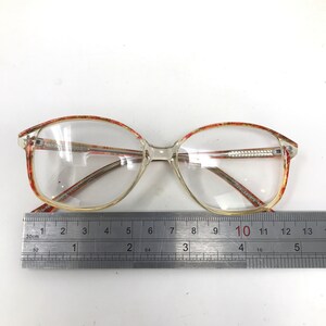 May include: Clear eyeglasses with a rectangular shape and a patterned red and brown design on the upper frame. The glasses are measured with a ruler, showing a length of approximately 5 inches.