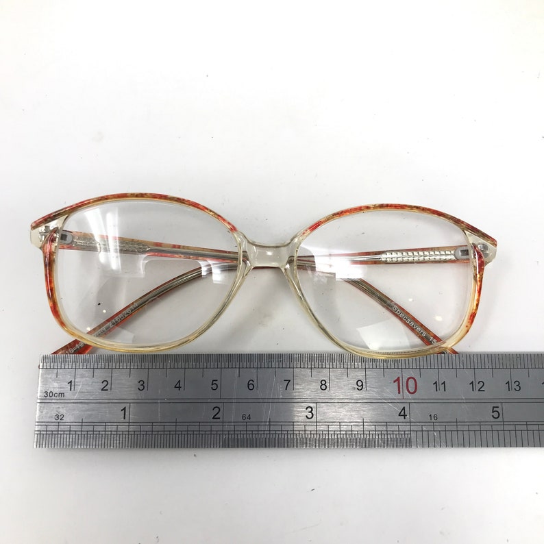 May include: Clear eyeglasses with a rectangular shape and a patterned red and brown design on the upper frame. The glasses are measured with a ruler, showing a length of approximately 5 inches.