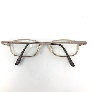 May include: Silver-toned metal eyeglasses with rectangular lenses. The frames have a thin, delicate design with dark brown temples. The glasses are positioned on a white surface, showcasing their minimalist aesthetic.