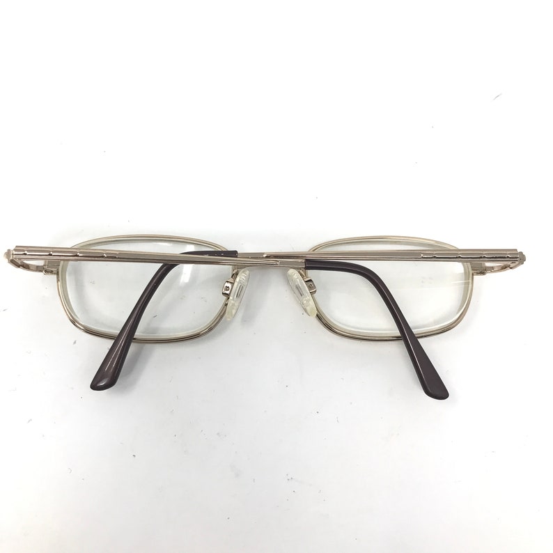 May include: Silver-toned metal eyeglasses with rectangular lenses. The frames have a thin, delicate design with dark brown temples. The glasses are positioned on a white surface, showcasing their minimalist aesthetic.