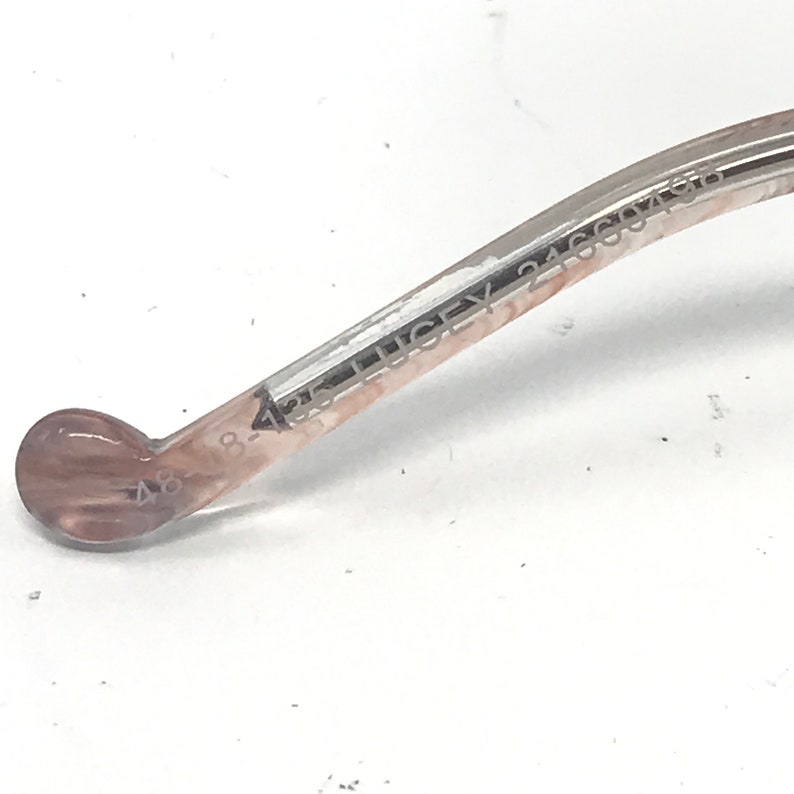 May include: Close-up of a section of eyeglasses. The frame is a light brown colour with the text "LUCKEY 24660496" and "48-4" printed on the arm. The end of the arm has a rounded, light brown tip.