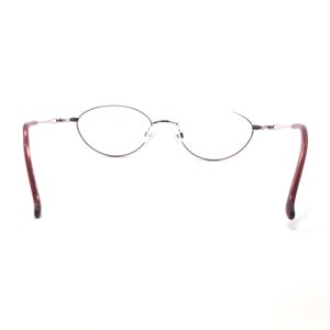 Puede incluir: Gafas de forma ovalada con montura fina y oscura y lentes transparentes. Las patillas son de color burdeos oscuro. Las gafas est&aacute;n sobre un fondo blanco.