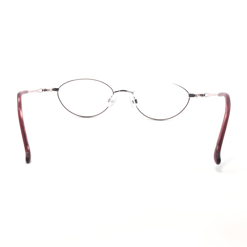 Puede incluir: Gafas de forma ovalada con montura fina y oscura y lentes transparentes. Las patillas son de color burdeos oscuro. Las gafas est&aacute;n sobre un fondo blanco.
