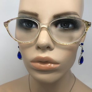 May include: Clear eyeglasses with a cat-eye shape and gold flecks on the frame. The glasses are displayed on a mannequin head, paired with dangling blue and silver earrings. The overall aesthetic is vintage and elegant.
