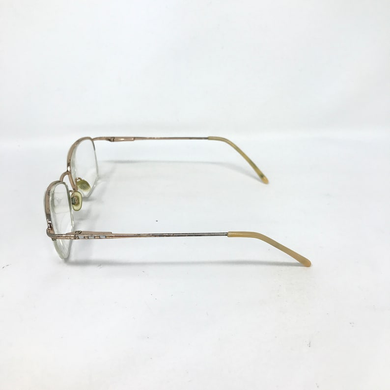 May include: A pair of eyeglasses with a gold-colored metal frame. The lenses are clear, and the temples have a light brown plastic coating. The glasses are set against a white background.