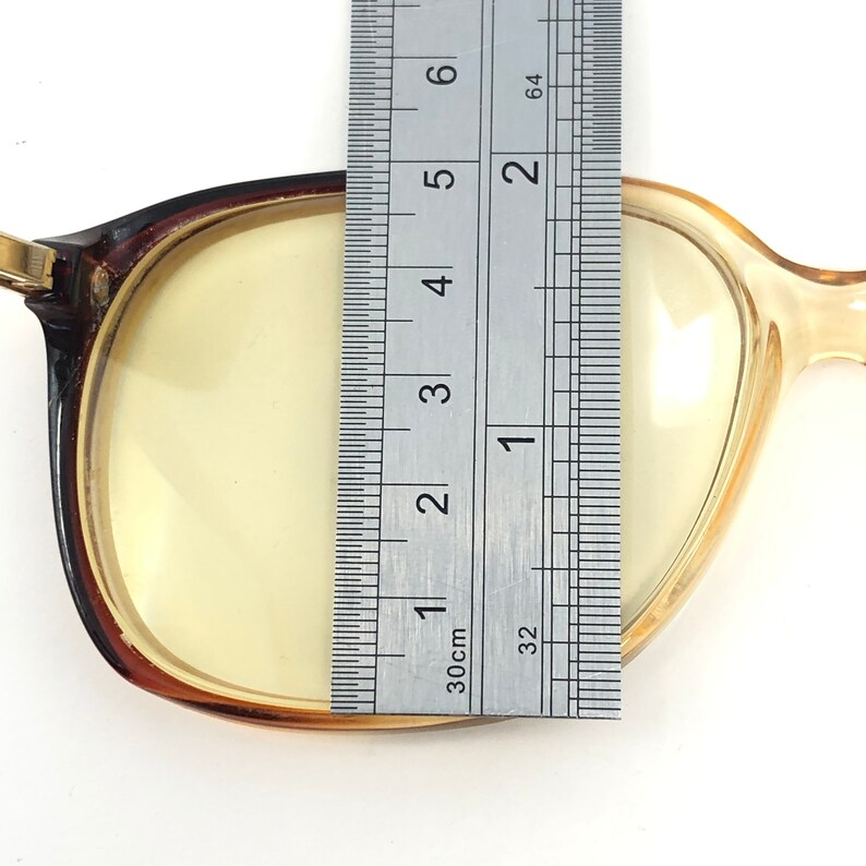 May include: A pair of eyeglasses with a brown frame and yellow lenses. A ruler is placed over the lens, measuring approximately 2 inches across. The glasses have a classic, rectangular shape.