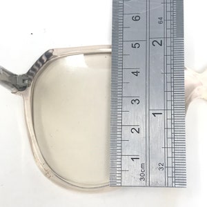 May include: Clear eyeglasses with a silver frame are measured with a ruler. The ruler shows the glasses are approximately 2 inches in height. The glasses have a rectangular shape and are set against a white background.
