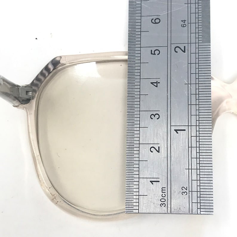May include: Clear eyeglasses with a silver frame are measured with a ruler. The ruler shows the glasses are approximately 2 inches in height. The glasses have a rectangular shape and are set against a white background.