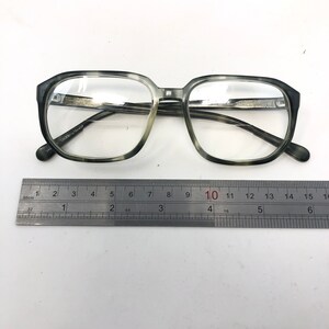 May include: Square-framed eyeglasses with a gray and white marbled pattern. The glasses are placed next to a metal ruler. The frame is thick and the lenses are clear. The ruler shows measurements in inches.