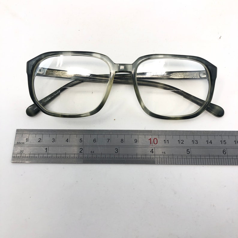 May include: Square-framed eyeglasses with a gray and white marbled pattern. The glasses are placed next to a metal ruler. The frame is thick and the lenses are clear. The ruler shows measurements in inches.