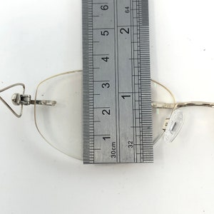 May include: A pair of rimless eyeglasses with a silver metal frame. The lens is measured with a ruler, showing a width of approximately 1.5 inches. The glasses have clear lenses and nose pads.