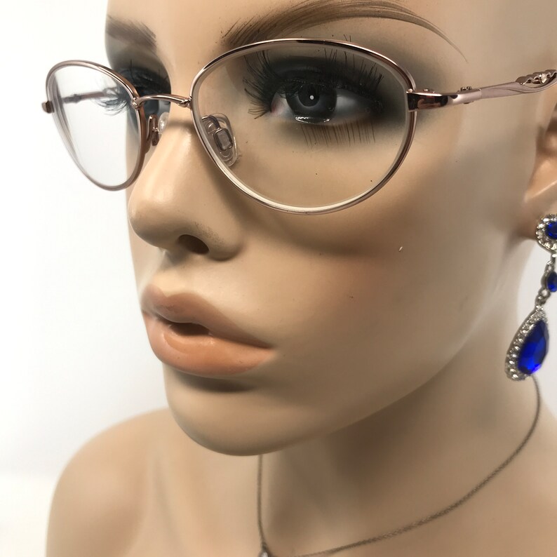 May include: Rose gold oval eyeglasses with clear lenses. The glasses have a delicate frame and are displayed on a mannequin. The mannequin also has long eyelashes and blue gemstone earrings.