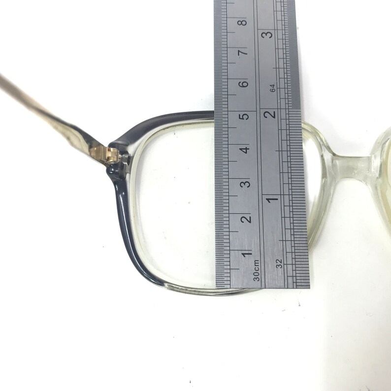 Pu&ograve; includere: Occhiali con montatura trasparente e bordo grigio scuro, misurati con un righello. La montatura &egrave; larga circa 5 cm. Gli occhiali hanno aste color oro e una forma rettangolare classica.